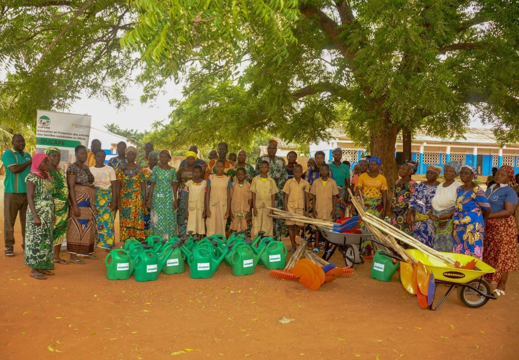 Photo de groupe élève en kaki et adultes accompagnés de matériels agricoles dans une cours d'école primaire.